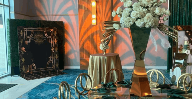 Great Gatsby themed party setup with custom gobo lighting projecting Art Deco fan patterns in warm orange tones on the walls, gold circular chairs, emerald green velvet cushions, a glass dining table, and a tall white floral centerpiece