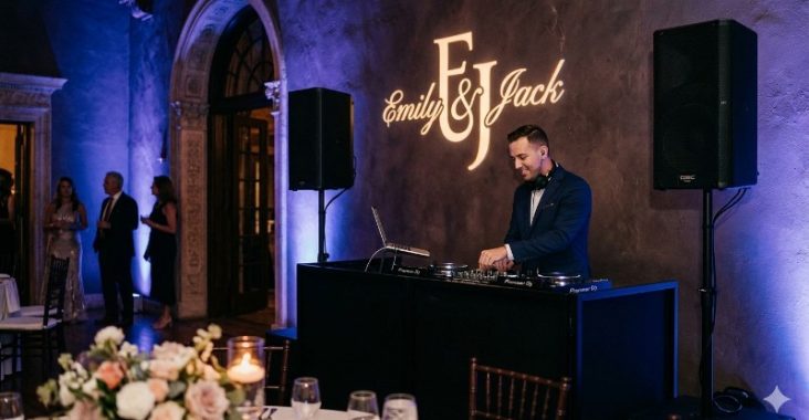 Custom wedding gobo projecting Emily and Jack monogram onto hardwood dance floor at elegant reception with soft warm ambient lighting, gold Chiavari chairs, and white tablecloths in background
